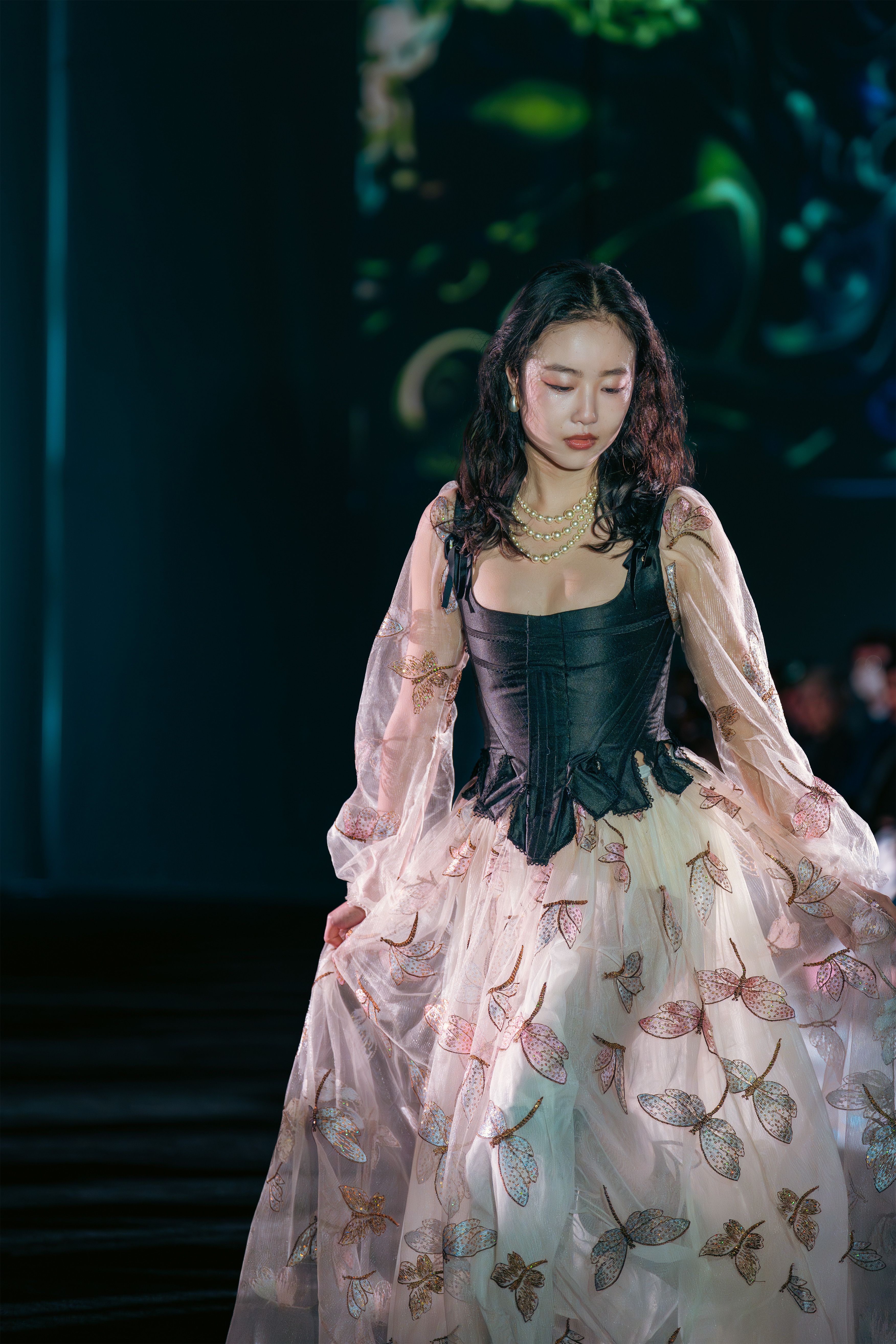 Model wears a black corset with a voluminous sheer overlay skirt embroidered with delicate multicolored butterflies. Her look is styled with soft waves and layered pearl necklaces.